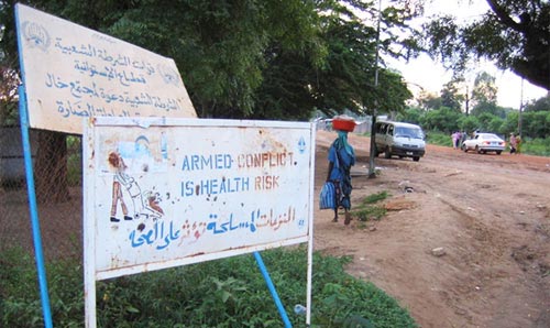 'Armed conflict' warning sign in foreground, and people going about daily business in the background.